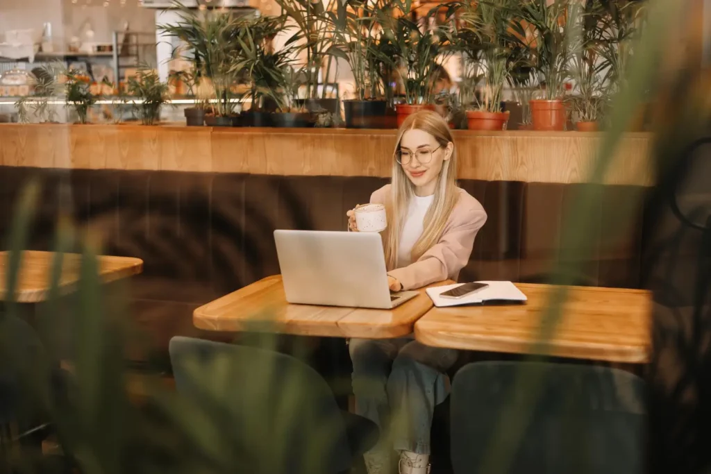 Estilista em uma cafeteria cheia de plantas, com um notebook, conferindo conteúdos na internet como blogs de moda.