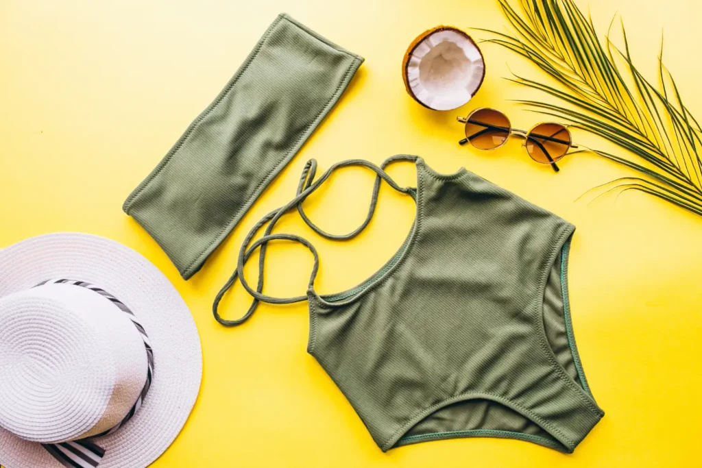 Biquíni verde de moda praia em um fundo amarelo com elementos como chapéu, folha, óculos de sol e metade de um coco.