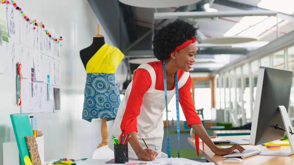 Diseñadora concentrada trabajando en su computadora, enfocada en el aumento de producción de su colección de moda, con bocetos y telas al fondo.