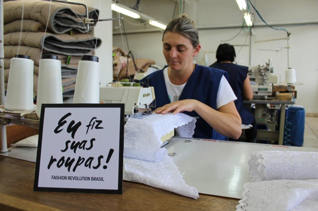 The image shows a woman working in a clothing company. In front of her, there's a sign with the message "I made your clothes", in Portuguese.