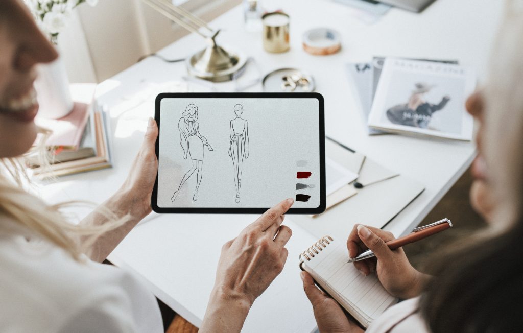 Croqui digital de moda desenhado em tablet e duas mulheres observando.