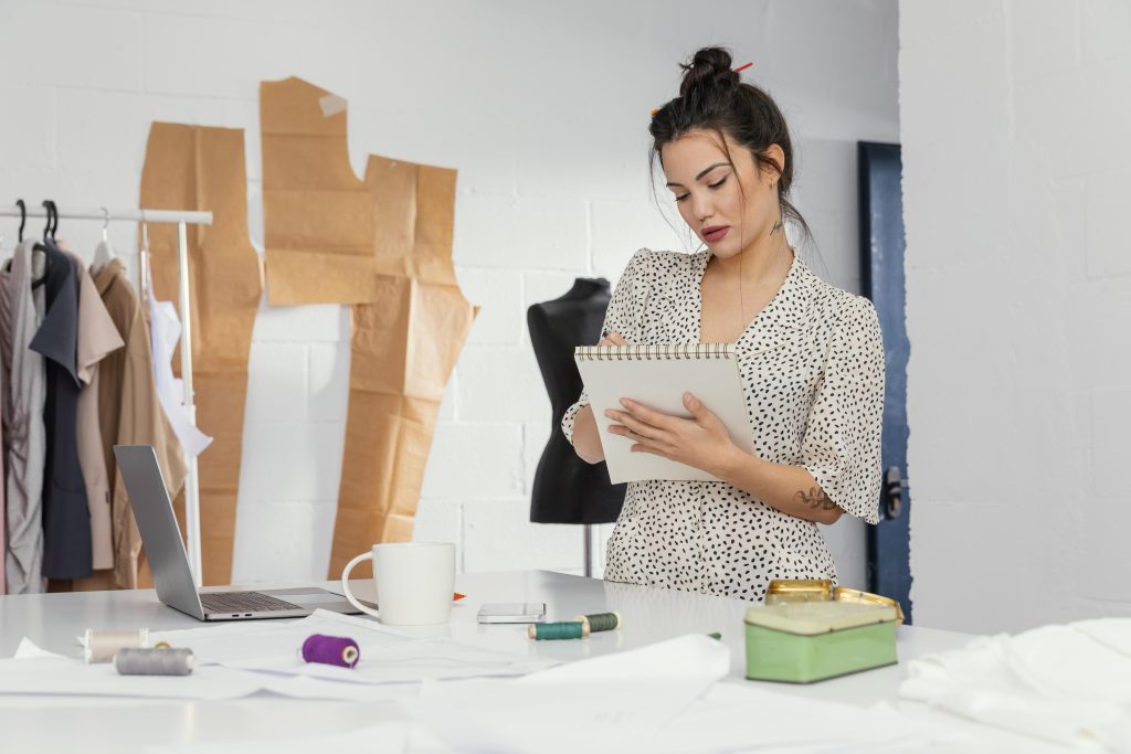 Mulher anotando em bloco de notas em local com moldes feitos em papel e algumas peças de roupa 