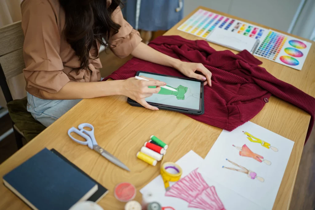 Mulher desenhando peça de roupa em tablet e ao lado acessórios de costura 