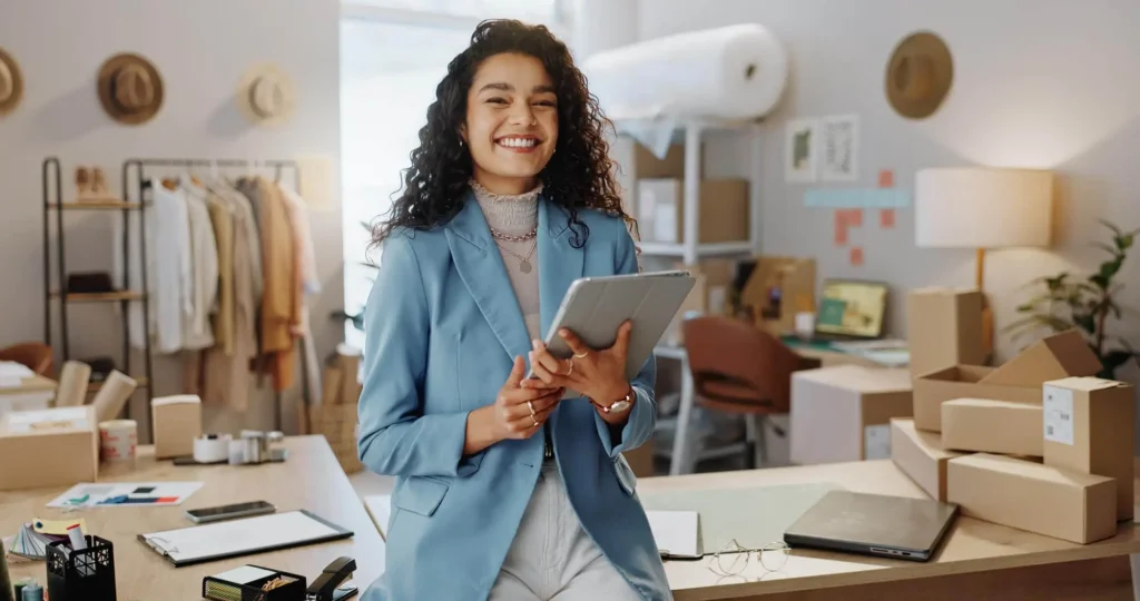 Mulher sorridente segurando um tablet em meio a materiais num atelier de moda.