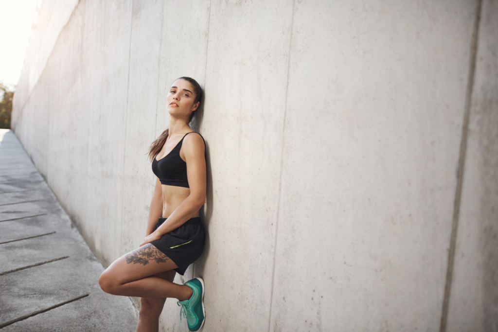 Mulher com roupas de corrida, com tecido esportivo, escorada numa parede.