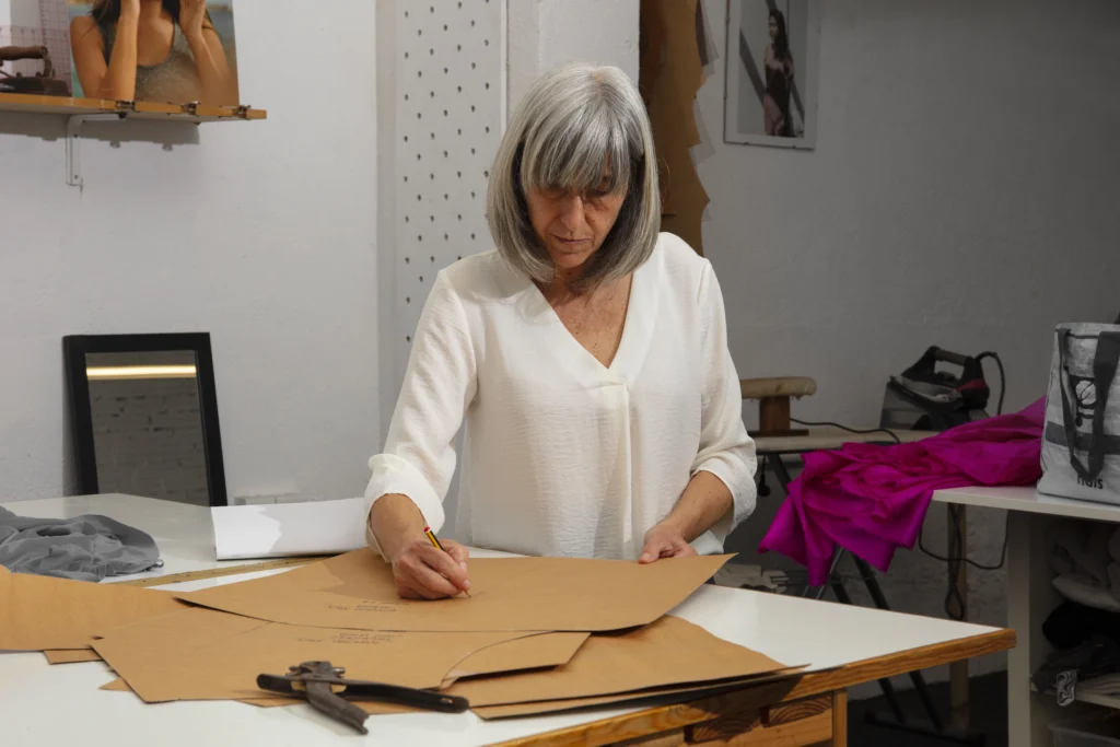 Una persona observando un patrón de costura en una mesa, mientras toma notas sobre cómo interpretar un patrón de costura y rodeada de herramientas y telas