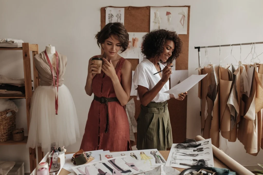 Donne fashion designer al lavoro in ufficio, una con caffè, l'altra con disegno in mano che studiano per la prossima creazione di moda