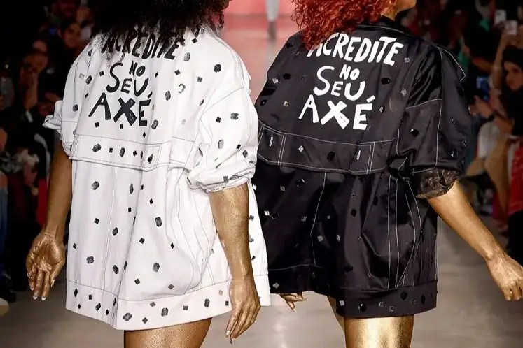 Modelos em desfile com uma jaqueta branca e uma jaqueta preta, ambas com a estampa escrita "Acredite no Seu Axé" nas costas. Créditos: Agência Fotosite/Divulgação/Revista Claudia