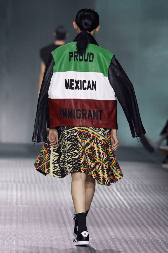 diseñadores de moda mexicanos: modelo desfilando con una chaqueta con la frase "Proud Mexican Immigrant" y los colores verde, blanco y rojo del diseñador Ricardo Seco.