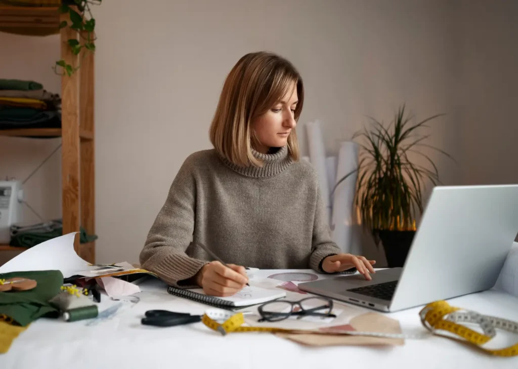 Estilista usando um computador, rodeada de materiais como tecidos, tesouras e pap&eacute;is.